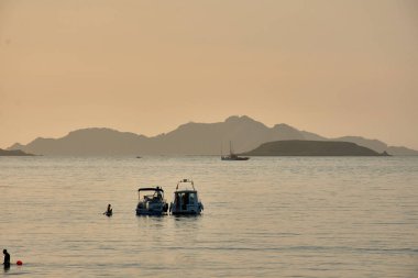 Güneş Baiona Körfezi 'nde batarken pruvaları görkemli Cies Adaları' na dönük tekneler sakin ve güzel bir manzara yaratıyor. Günbatımının ılık, altın rengi sakin sulardan yansıyarak deniz yolculuğunun güzelliğini arttırır.