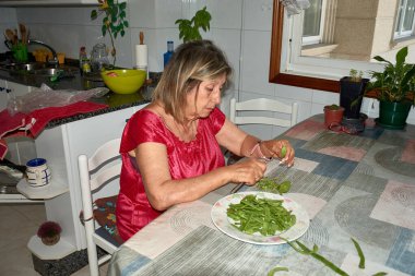 Mutfak masasında oturan yaşlı bir kadın dikkatlice yeşil fasulyeleri bıçakla kesiyor. Sahne, geleneksel yemek pişirme yöntemlerinin nesilden nesile aktarıldığı günlük hayatın basitliğini ve konforunu yansıtıyor..