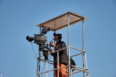 Bayona,Pontevedra,Spain; August,27,2024;a television cameraman positioned on an elevated platform, capturing live footage of a sporting event. The camera operator, equipped with a high-end camera, focuses intensely on the action below, ensuring every clipart