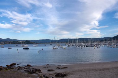 İspanya, Pontevedra, Bayona 'daki Barbeira Sahili' nden sakin bir sahne. Körfezin sakin suları, yumuşak, ince bulutlu, berrak mavi bir gökyüzünün altında barış içinde demirlemiş yelkenli ve küçük yatlarla doludur..