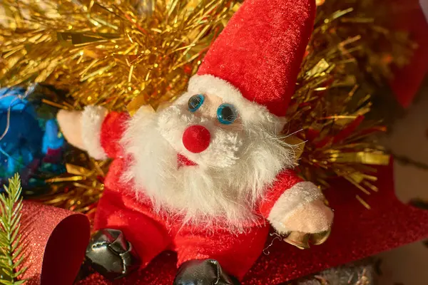 Noel Baba şenlikli bir Noel sahnesinin ortasında oturan pelüş oyuncağı. Noel Baba figürü, tüylü beyaz sakalı, parlak kırmızı elbisesi ve mavi gözleriyle, tatil atmosferini güzelleştiren parıltılı altın bir arkaplan ile çevrilidir..