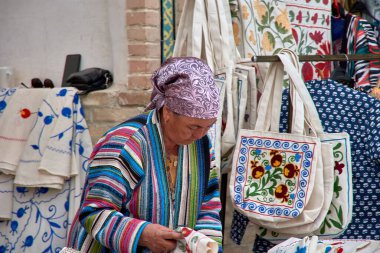 Buhara, Özbekistan; Eylül 19.2024: Özbekistan 'ın canlı Buhara kentindeki meraklı turistlere zarif ipek parçaları ustaca satan yaşlı bir kadın. Sıcak gülüşü ve ipek kumaşların karmaşık desenleri.