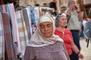 Khiva, Özbekistan; 21 Eylül 2024: Özbekistan 'ın Khiva kentindeki pazarlarda tekstil standında çalışan yaşlı bir Özbek kadın. Geleneksel bir elbise ve desenli bir başörtüsü giyerek renkli kumaşları dikkatlice düzenler.