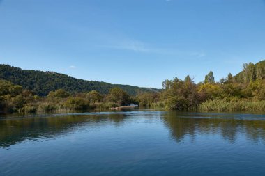 Hırvatistan 'daki Krka Ulusal Parkı' nda huzurlu bir tekne yolculuğu. Teknenin dalgalanması berrak sularda dalgalanır, etrafındaki yemyeşil tepeleri yansıtır ve parlak mavi gökyüzü tüylü beyaz bulutlarla beneklenir.. 