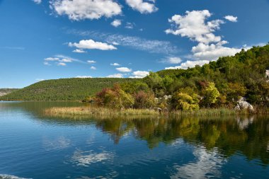 Hırvatistan 'daki Krka Ulusal Parkı' nda huzurlu bir tekne yolculuğu. Teknenin dalgalanması berrak sularda dalgalanır, etrafındaki yemyeşil tepeleri yansıtır ve parlak mavi gökyüzü tüylü beyaz bulutlarla beneklenir..