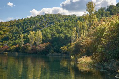 Hırvatistan 'daki Krka Ulusal Parkı' nda huzurlu bir tekne yolculuğu. Teknenin dalgalanması berrak sularda dalgalanır, etrafındaki yemyeşil tepeleri yansıtır ve parlak mavi gökyüzü tüylü beyaz bulutlarla beneklenir.. 