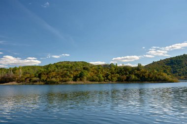 Hırvatistan 'daki Krka Ulusal Parkı' nda huzurlu bir tekne yolculuğu. Teknenin dalgalanması berrak sularda dalgalanır, etrafındaki yemyeşil tepeleri yansıtır ve parlak mavi gökyüzü tüylü beyaz bulutlarla beneklenir.. 