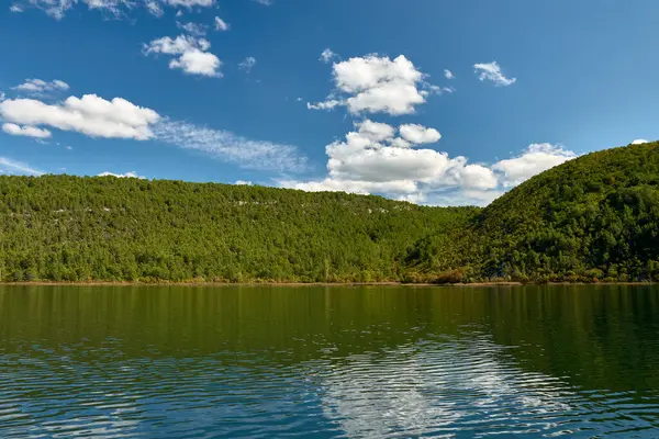 Hırvatistan 'daki Krka Ulusal Parkı' nda huzurlu bir tekne yolculuğu. Teknenin dalgalanması berrak sularda dalgalanır, etrafındaki yemyeşil tepeleri yansıtır ve parlak mavi gökyüzü tüylü beyaz bulutlarla beneklenir.. 