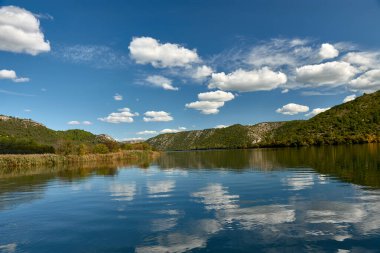 Hırvatistan 'daki Krka Ulusal Parkı' nda huzurlu bir tekne yolculuğu. Teknenin dalgalanması berrak sularda dalgalanır, etrafındaki yemyeşil tepeleri yansıtır ve parlak mavi gökyüzü tüylü beyaz bulutlarla beneklenir.
