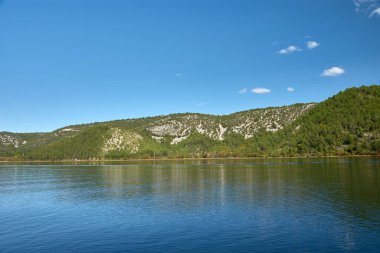 Hırvatistan 'daki Krka Ulusal Parkı' nda huzurlu bir tekne yolculuğu. Teknenin dalgalanması berrak sularda dalgalanır, etrafındaki yemyeşil tepeleri yansıtır ve parlak mavi gökyüzü tüylü beyaz bulutlarla beneklenir.. 