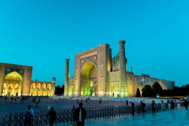 SAMARKAND, UZBEKISTAN - 17 Eylül 2024: Semerkant, Özbekistan 'daki Registan Meydanı, akşam gökyüzünün altında aydınlandı. Tarihi İpek Yolu boyunca önemli bir konum olarak Registan Meydanı üç büyük medresesi ile ünlüdür.