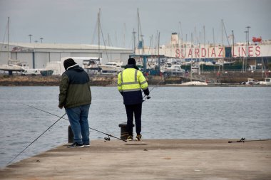 Vigo, Pontevedra, İspanya; Aralık, 28.2024: Vigos Orillamar limanının kenarında duran balıkçılar oltasını sakin sulara atıyorlar. Kenetlenmiş balıkçı tekneleri ve hareketli bir deniz zeminiyle çevrili.