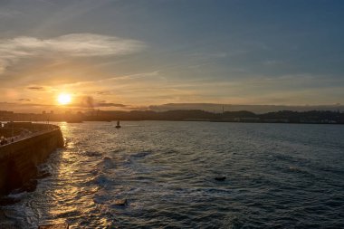 Görüntü, güneşin ufka doğru zarifçe alçaldığını gösteriyor. Denizin engin genişliğinde ılık bir altın parıltı yayıyor. Gökyüzü canlı portakallar ve kırmızılardan yumuşak pembelere ve morlara kadar uzanan bir renk senfonisi ile alevler içinde.