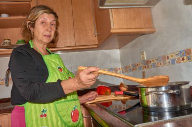 Bir büyükanne şık ve modern bir mutfakta yemek pişirirken görülüyor. Önlük takarak, taze malzemelerle birlikte dikkatlice bir yemek hazırlıyor. Görüntü, ev yemeklerinin sıcaklığını, geleneği modernlikle harmanlamasını vurguluyor. Aile teması için mükemmel.,