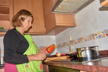 Bir büyükanne şık ve modern bir mutfakta yemek pişirirken görülüyor. Önlük takarak, taze malzemelerle birlikte dikkatlice bir yemek hazırlıyor. Görüntü, ev yemeklerinin sıcaklığını, geleneği modernlikle harmanlamasını vurguluyor. Aile teması için mükemmel.,