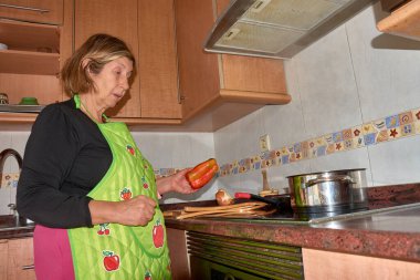 Bir büyükanne şık ve modern bir mutfakta yemek pişirirken görülüyor. Önlük takarak, taze malzemelerle birlikte dikkatlice bir yemek hazırlıyor. Görüntü, ev yemeklerinin sıcaklığını, geleneği modernlikle harmanlamasını vurguluyor. Aile teması için mükemmel.,