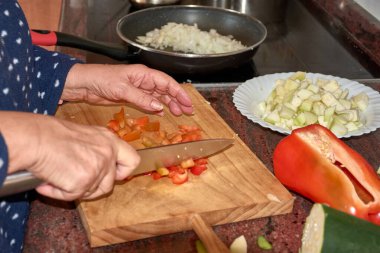 Yaşlı ellerin tahta kesme tahtasındaki keskin bir bıçakla taze sebzeleri dilimlediği yakın plan bir görüntü. Fotoğraf, geleneksel yemek pişirme sanatını sergileyerek, yemek hazırlamada özen ve hassasiyeti yakalıyor. Dekor rahat bir mutfak, mükemmel. 