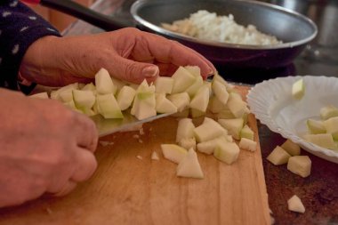 Yaşlı ellerin tahta kesme tahtasındaki keskin bir bıçakla taze sebzeleri dilimlediği yakın plan bir görüntü. Fotoğraf, geleneksel yemek pişirme sanatını sergileyerek, yemek hazırlamada özen ve hassasiyeti yakalıyor. Dekor rahat bir mutfak, mükemmel. 