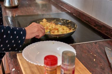 Yaşlı ellerin taze sebzeleri dilimleyip soba tepsisine koyduğu yakın plan bir görüntü. Sahne, sıcacık bir mutfakta ev yapımı bir saut hazırlamayı gösteriyor. Geleneklere, sağlıklı yemeklere ve aile odaklı yaşam tarzlarına önem veriliyor.