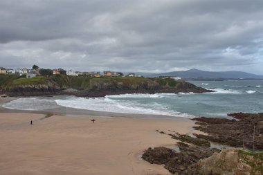 Tapia de Casariego, Asturias, İspanya; 29 Mart 2025: Tapia de Casariego, İspanya 'da Aguileiro Sahili boyunca kırmızı bir sörf tahtası taşıyan yalnız bir sörfçü. Engebeli kıyı şeridi, güçlü dalgalar ve karamsar gökyüzü dramatik bir sahne yaratır.. 