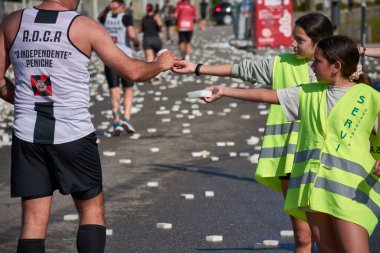 Nigran, Pontevedra, İspanya; 6 Nisan 2025: Yüksek görüşlü yelek giyen genç gönüllüler, İspanya 'nın Nigrn kentindeki Vig Bay Yarı Maratonu sırasında koşuculara suya batırılmış süngerler dağıtıyor. 