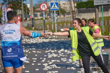 Nigran, Pontevedra, İspanya; 6 Nisan 2025: Yüksek görüşlü yelek giyen genç gönüllüler, İspanya 'nın Nigrn kentindeki Vig Bay Yarı Maratonu sırasında koşuculara suya batırılmış süngerler dağıtıyor. 