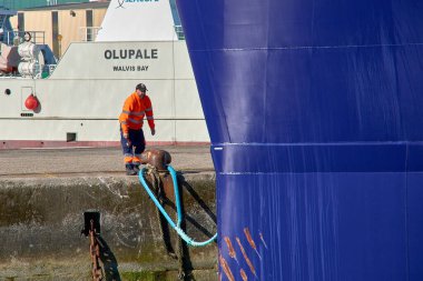 Vigo, Pontevedra, İspanya; 6 Mayıs 2025: Vigo 'da bir rıhtıma demirlemiş büyük, parlak mavi bir geminin demirleme çizgilerini manipüle eden yüksek görüşlü giysiler giyen bir liman işçisi. 