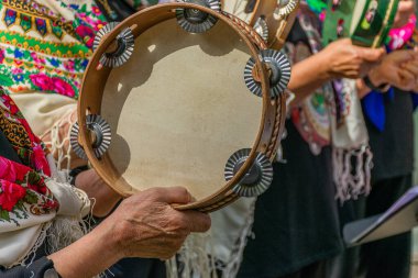 Buruşuk, bilge ellerin ayrıntılı yakın çekimi. Soluk kafaları ve metal şıngırtılarıyla ahşap bir tefi nazikçe tutuyor ve çalıyorlar. Çiçekler ve nakışlarla bezenmiş Galiçya kantarezlerinin renkli geleneksel kostümleri.