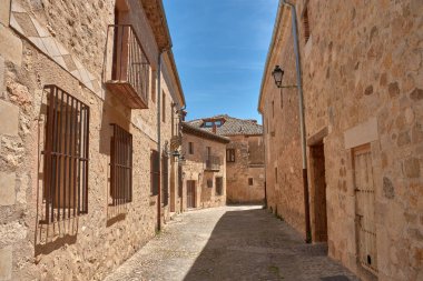 Huzurlu bir kaldırım taşı sokak, demirden yapılma balkonları ve parmaklıklı pencereleri olan antik taş evler tarafından çevrili, canlı mavi gökyüzü altında hafif kıvrımlar, Pedraza 'nın geleneksel mimarisine bir bakış sunuyor.