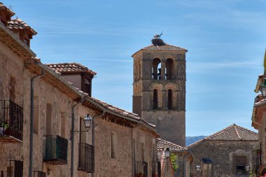 Pedraza 'nın kilisesinin görkemli taş çan kulesi etrafındaki evlerin kiremitli çatılarının üzerinde belirgin bir şekilde yükselir, açık mavi gökyüzünün altında bir leylek yuvası bulunur.. 