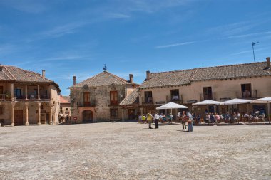 Pedraza, Segovia, İspanya; 24 Mayıs 2025: Pedraza 'nın kaldırım taşlarıyla döşenmiş ikonik Plaza Belediye Başkanı, ahşap balkonlu geleneksel taş evlerle çevrili, açık mavi gökyüzü altında güneşli terasların keyfini çıkaran insanlarla dolup taşıyor.. 