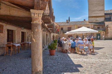Pedraza, Segovia, İspanya; 24 Mayıs 2025: Pedraza 'nın kaldırım taşlarıyla döşenmiş ikonik Plaza Belediye Başkanı, ahşap balkonlu geleneksel taş evlerle çevrili, açık mavi gökyüzü altında güneşli terasların keyfini çıkaran insanlarla dolup taşıyor.. 