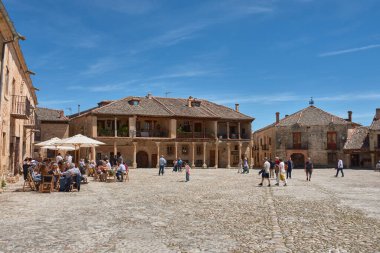 Pedraza, Segovia, İspanya; 24 Mayıs 2025: Pedraza 'nın kaldırım taşlarıyla döşenmiş ikonik Plaza Belediye Başkanı, ahşap balkonlu geleneksel taş evlerle çevrili, açık mavi gökyüzü altında güneşli terasların keyfini çıkaran insanlarla dolup taşıyor.. 