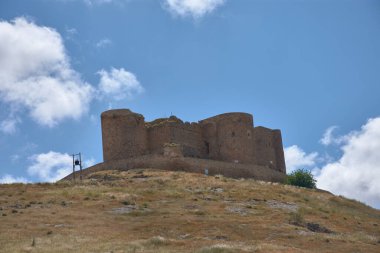 Görkemli bir ortaçağ şatosu dağınık bulutlarla dolu açık mavi bir gökyüzünün altında kuru, çimenli bir tepeyi taçlandırır. Antik taşın toprak tonları canlı gökyüzü ile zıt, miras ve gücü çağrıştıran; seyahat, tarih veya kültürel temalar için ideal.