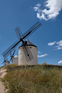 Siyah bıçakları olan ikonik beyaz yel değirmenleri, altın otlar ve kayalık arazilerle çevrili, canlı mavi gökyüzünün altında bir tepe boyunca uzanır. Kompozisyon geleneği, sakinliği ve kültürel zenginliği aktarıyor.