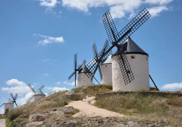 Siyah bıçakları olan ikonik beyaz yel değirmenleri, altın otlar ve kayalık arazilerle çevrili, canlı mavi gökyüzünün altında bir tepe boyunca uzanır. Kompozisyon geleneği, sakinliği ve kültürel zenginliği aktarıyor.