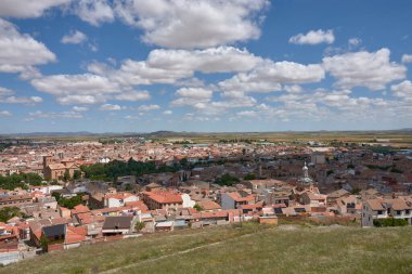 Consuegra, Toledo 'nun geniş açılı bir manzarası, kırmızı kiremitli çatılar, tarihi bir kilise ve dağınık bulutlarla kaplı mavi gökyüzünün altındaki tarım arazilerini gösteriyor. Sıcak melodiler ve açık mekan huzurlu bir ortam oluşturuyor.