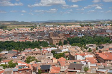Consuegra, Toledo 'nun geniş açılı bir manzarası, kırmızı kiremitli çatılar, tarihi bir kilise ve dağınık bulutlarla kaplı mavi gökyüzünün altındaki tarım arazilerini gösteriyor. Sıcak melodiler ve açık mekan huzurlu bir ortam oluşturuyor.