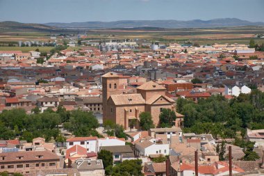 Consuegra, Toledo 'nun geniş açılı bir manzarası, kırmızı kiremitli çatılar, tarihi bir kilise ve dağınık bulutlarla kaplı mavi gökyüzünün altındaki tarım arazilerini gösteriyor. Sıcak melodiler ve açık mekan huzurlu bir ortam oluşturuyor.