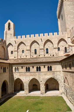 Avignon, Provence, Fransa 'daki Palais des Papes' in canlı ve bulutsuz mavi gökyüzünün altındaki avlu ve iç binalarının manzarası. Kompozisyon, birden fazla kemerle süslenmiş taş duvarların sağlamlığını vurguluyor. 