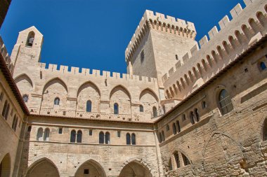 Avignon, Provence, Fransa 'daki Palais des Papes' in canlı ve bulutsuz mavi gökyüzünün altındaki avlu ve iç binalarının manzarası. Kompozisyon, birden fazla kemerle süslenmiş taş duvarların sağlamlığını vurguluyor. 