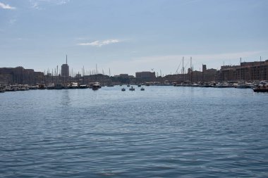 Yelkenliler ve yatlar mavi gökyüzünün altında Marsilya limanında hizalanırken, kent modern binalar ve tarihi kubbelerle arka planda yayılırken, direkleri sakin sularda yansıyor.. 