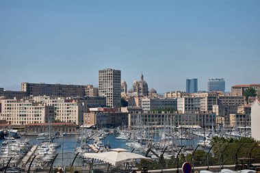 Yelkenliler ve yatlar mavi gökyüzünün altında Marsilya limanında hizalanırken, kent modern binalar ve tarihi kubbelerle arka planda yayılırken, direkleri sakin sularda yansıyor.. 