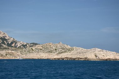 Hareket eden bir teknenin kıç tarafından Marsilya Calanques 'in heybetli kayalık silueti ufukta görünür. Akdeniz' in yoğun maviliği ve açık bir gökyüzü ile çevrilidir.. 