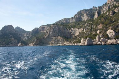 Hareket eden bir teknenin kıç tarafından Marsilya Calanques 'in heybetli kayalık silueti ufukta görünür. Akdeniz' in yoğun maviliği ve açık bir gökyüzü ile çevrilidir.. 