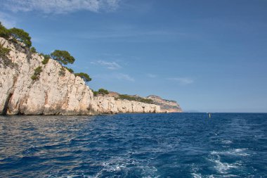 Hareket eden bir teknenin kıç tarafından Marsilya Calanques 'in heybetli kayalık silueti ufukta görünür. Akdeniz' in yoğun maviliği ve açık bir gökyüzü ile çevrilidir.. 