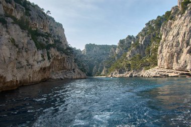 Hareket eden bir teknenin kıç tarafından Marsilya Calanques 'in heybetli kayalık silueti ufukta görünür. Akdeniz' in yoğun maviliği ve açık bir gökyüzü ile çevrilidir.. 