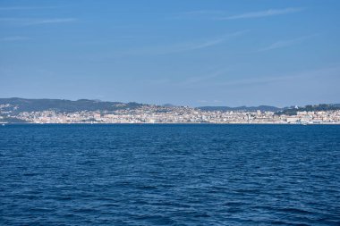 Vigo Estuary 'nin merkezinden kentsel gelişimi açık bir gökyüzünün altında kıyı boyunca uzanan geniş şehir manzarasının tadını çıkarabilirsiniz. Mavi deniz ön planda yer alır, parlak ve dengeli bir manzaraya derinlik ve kontrastlık sağlar.. 