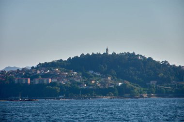 Vigo halklarının ortasındaki bir tekneden, La Guia Dağı sembolik yeşil bir koni gibi yükselerek, yapraklı ağaçlarla çevrelenmiş, şehrin ve Galiçya denizinin panoramik manzarasını sunar. Yoğun mavi renkler...