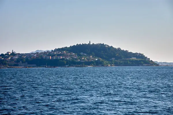 Vigo halklarının ortasındaki bir tekneden, La Guia Dağı sembolik yeşil bir koni gibi yükselerek, yapraklı ağaçlarla çevrelenmiş, şehrin ve Galiçya denizinin panoramik manzarasını sunar. Yoğun mavi renkler...